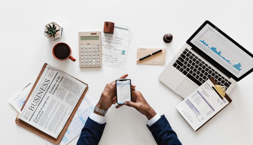 a person's hands holding a phone with other office items laid out around them on a white desk