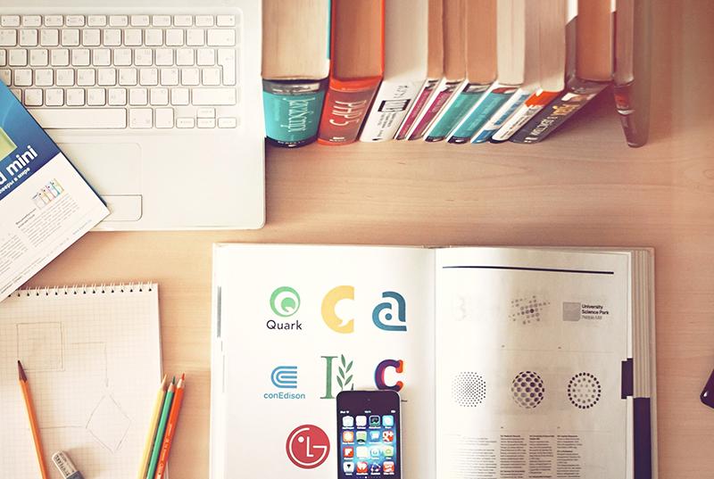 an open book with an iphone on top of it. the desk it's on also has a stack of books behind the open book and a laptop next to the stack