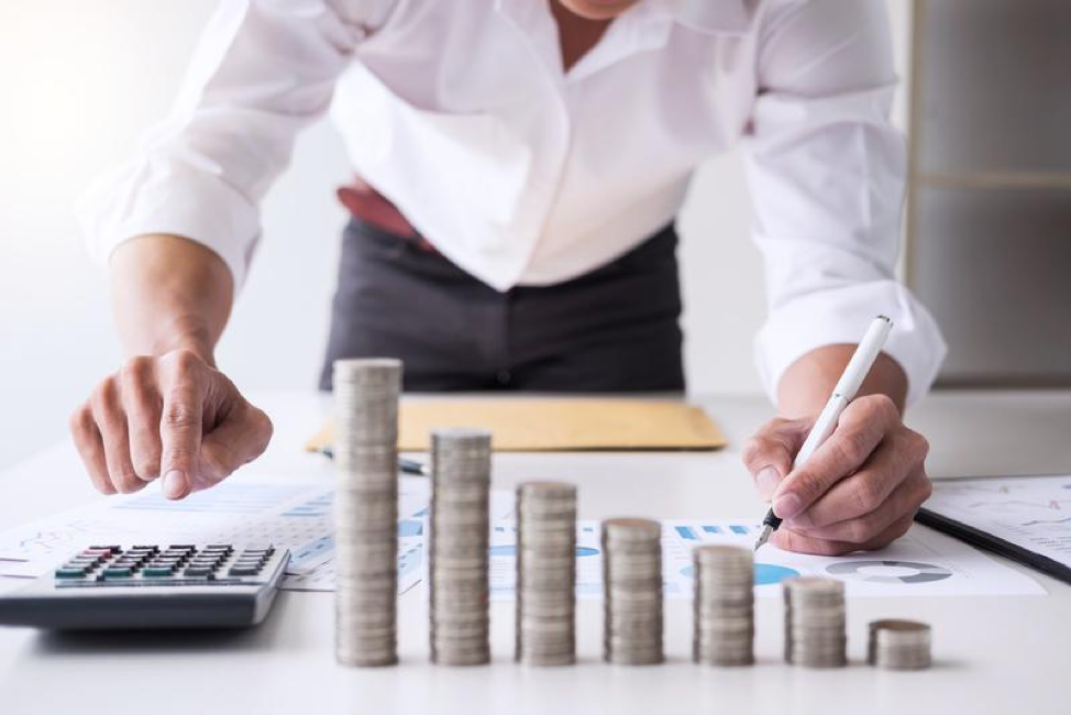 person hunched over a desk with a pen in their hand writing on a piece of paper. a stack of coins sits in front of them in ascending size.