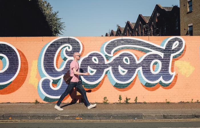 an orange wall on the side of a street. a person walks left to right in front of the wall on a sidewalk. the wall has dark blue graffitied typography that says "good"