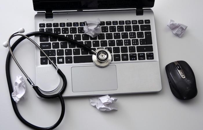 an open laptop with a stethoscope on top of it. a wireless mouse sits to the right of the laptop and crumpled white pieces of paper are around the laptop