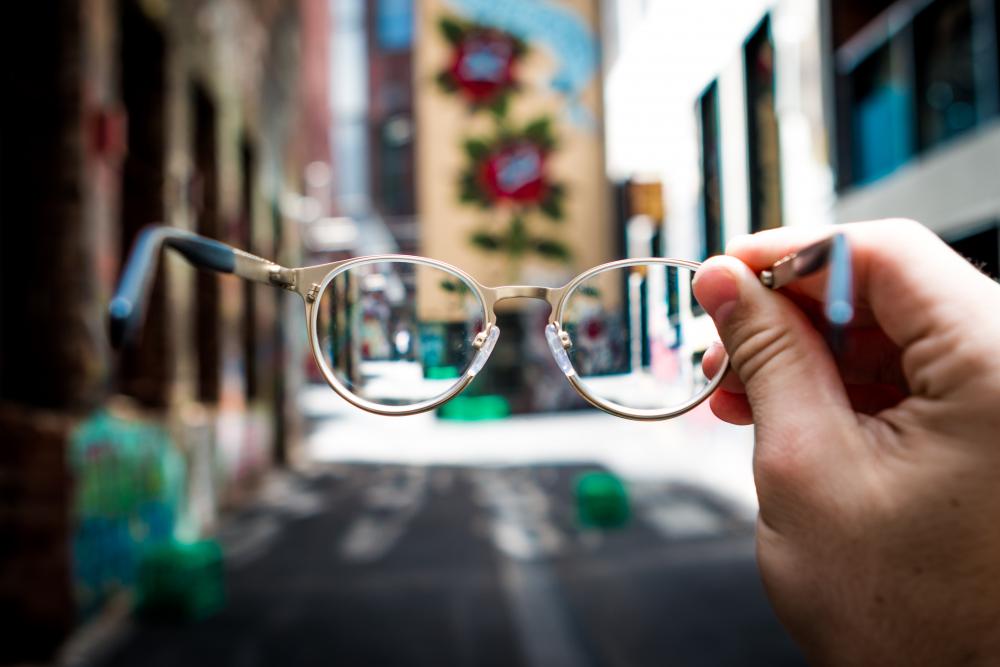 a hand holding out a pair of round glasses as the camera sees through them. the background is a city street