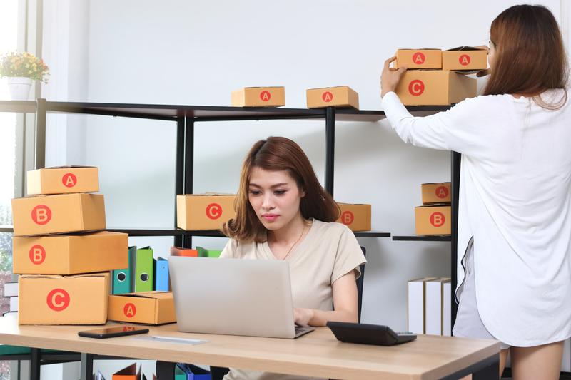 2 women in an office, one is sitting facing the viewer at a desk on a laptop, numerous boxes are on the desk and around the room. the 2nd woman's back is facing the viewer and she is adjusting one of the boxes behind the 1st woman