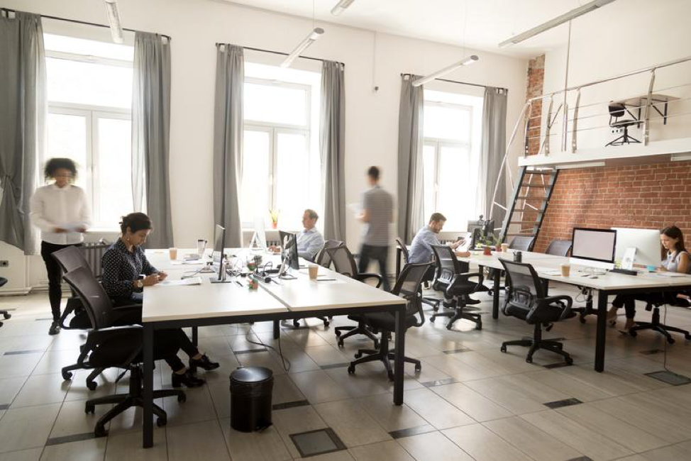 an office with many people in it sitting at desks working on computers or walking around. the office is very modern-looking and has wide floor-to-ceiling windows