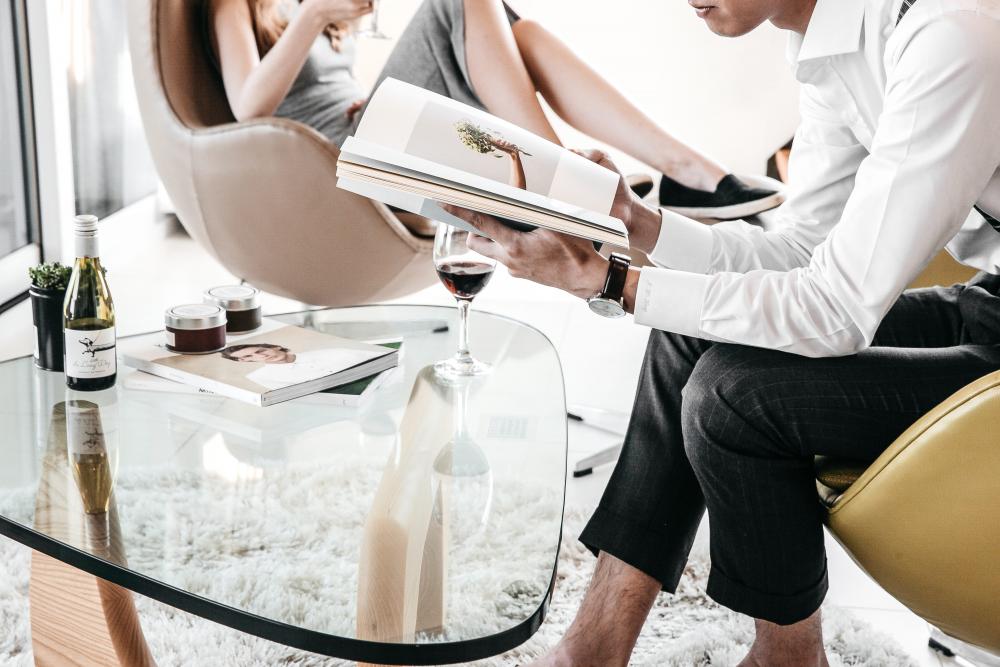 person sitting reading a book at a table with another person sitting further away in a chair also reading. both of their heads are out of the shot