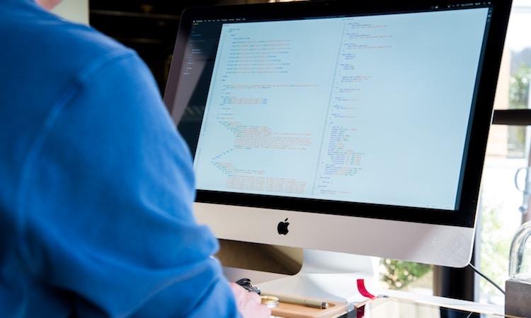 a person in a blue shirt sits in front of an imac computer that has code on the screen