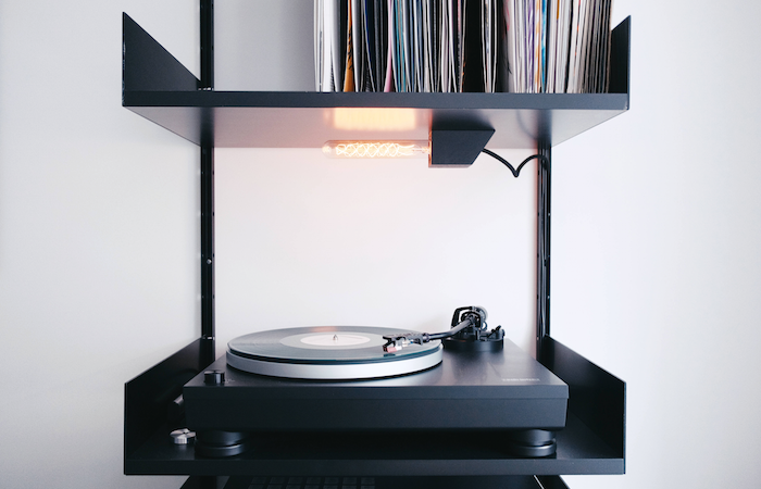 a record player with a record on it. the player sits on a piece of furniture that is the same width as the player, and has a top section with shelving that houses various records.