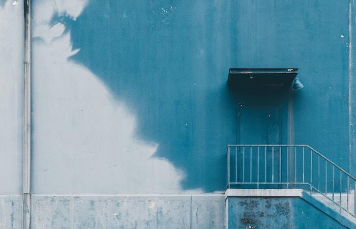 the side of a half blue painted building. there's a raised platform lining the building with a little railing and door on the right side