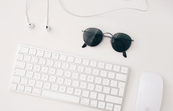 overhead image of a white keyboard, white mouse, white wired headphones, and a pair of black sunglasses