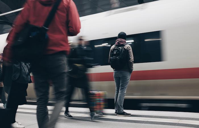 a person wearing a black backpack waits for a train as it whizzes by them. in the unfocused shot of the photo, 3 others wait for the train or walk past