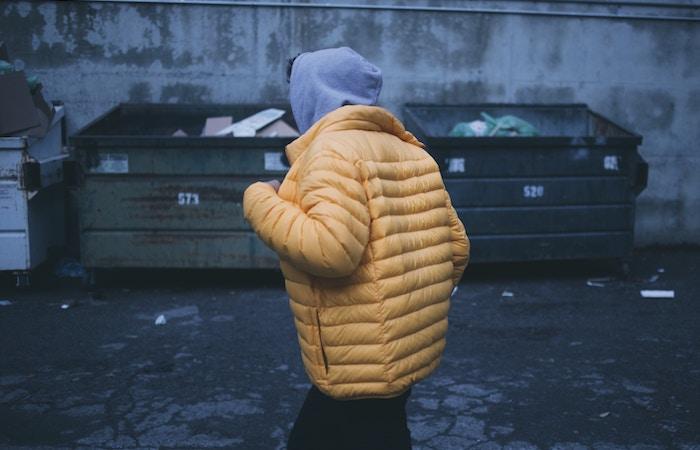 a person runs by 2 dumpsters wearing a grey hoodie and a yellow puff jacket